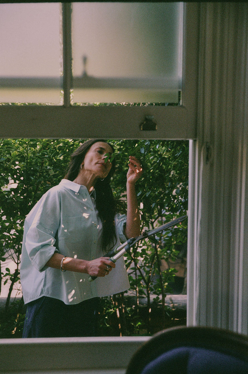 A woman is standing outside by tall green hedges, with the camera looking through an open sash window. She is holding hedge clippers and reaching for the leaves of the hedge, smelling them. She is wearing Saywood's Lela Colourblock Boxy Shirt in green and blue pastel, with her sleeves rolled up. Worn with the Amelia Wide Leg Trousers in Japanese Denim.