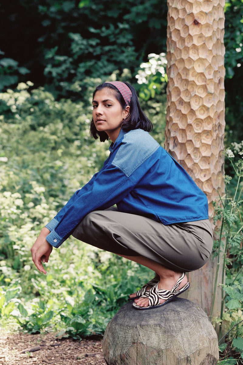 A woman squats on top of a round carved wooden bolder with a carved wooden post behind her, surrounded by greenery and nature, with her arms relaxed by her side, looking sideways. She wears Saywood's Japanese Denim Lela Patchwork Shirt, with light wash contrast cuffs, pockets and collar. Worn with the khaki Amelia Trousers and animal stripe monochrome sandals, with a red check headband in her hair. Shot from the side.